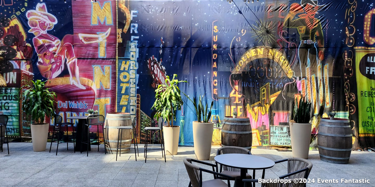 Outdoor seating area with tables and chairs, surrounded by plants in pots and barrels, set against a colorful backdrop depicting a vibrant, Las Vegas street scene with neon signs.