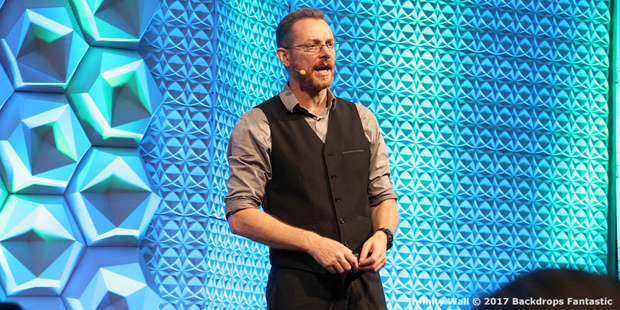 A man wearing glasses, a vest over a button-up shirt, speaks on stage with a geometric blue-green backdrop.