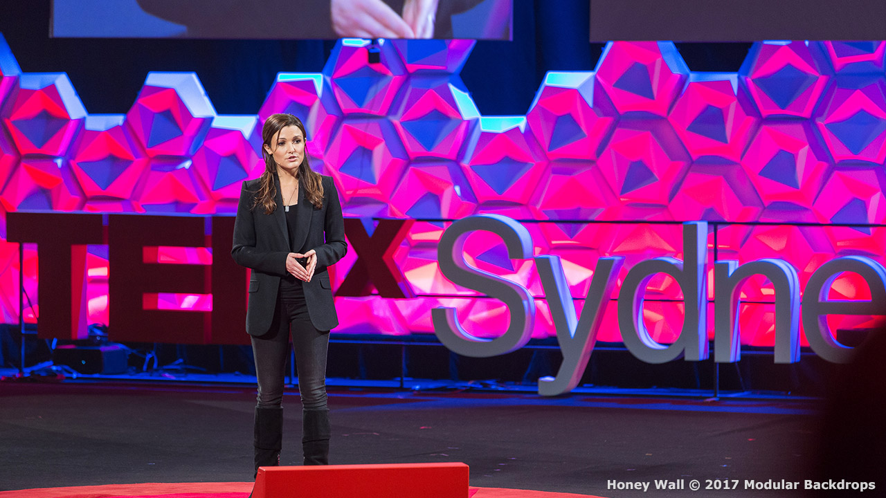 A person stands on stage giving a presentation at TEDxSydney with a geometric red and pink backdrop and the event logo displayed.