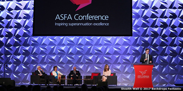 A panel of speakers sits on a stage in front of a geometric backdrop at the ASFA Conference, enhanced by Stealth Wall Modular Panels. A man stands at a podium speaking, with text "Inspiring superannuation excellence" displayed on the screen above.