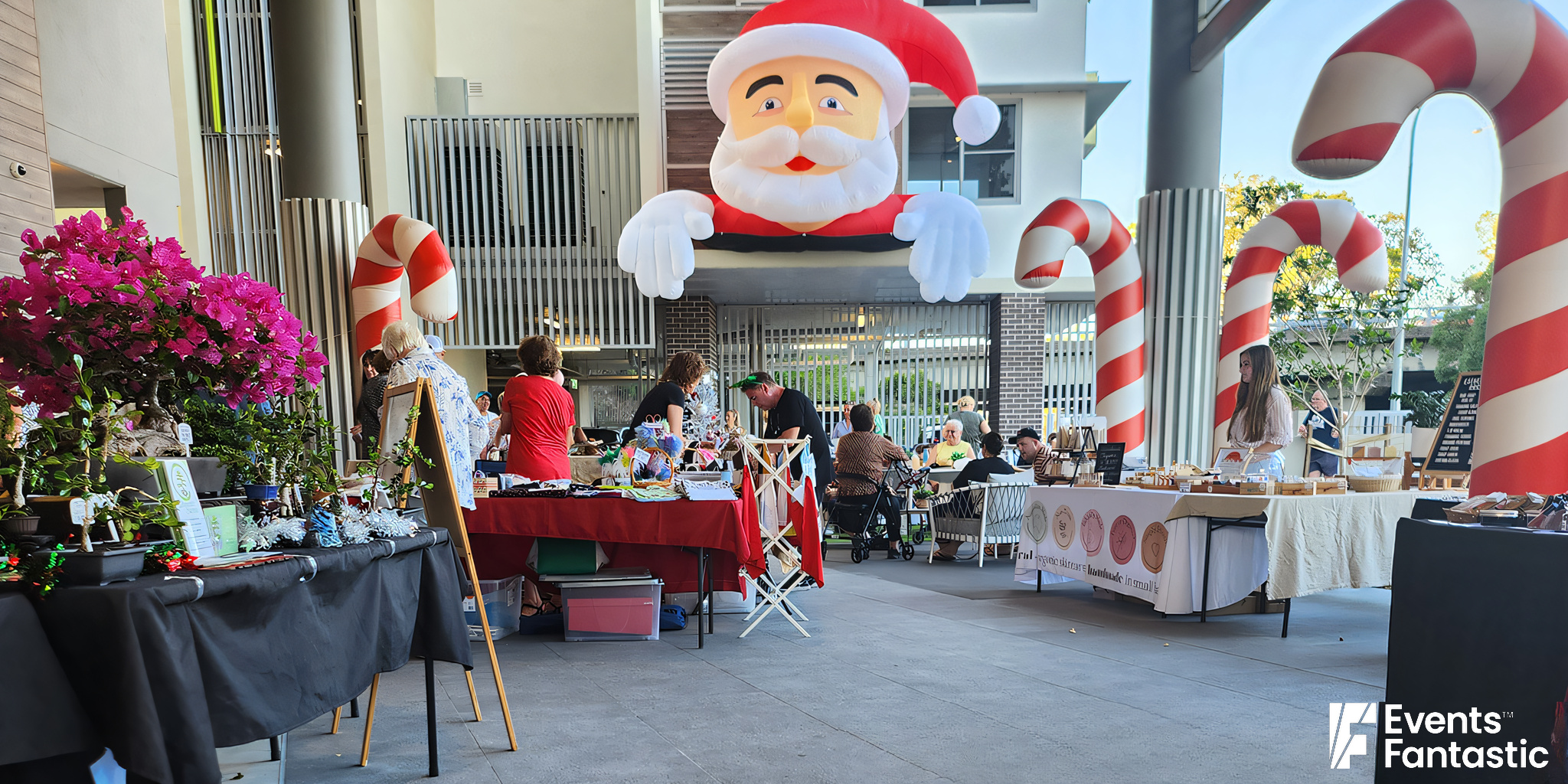 Outdoor holiday market with stalls displaying various items, a large inflatable Santa figure, and several inflatable candy cane decorations. People are browsing and interacting at the event.