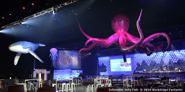 Large indoor event space with inflatable creatures, including a blue whale, purple octopus, and jellyfish suspended from the ceiling. Empty high-top tables and a bar are visible below.