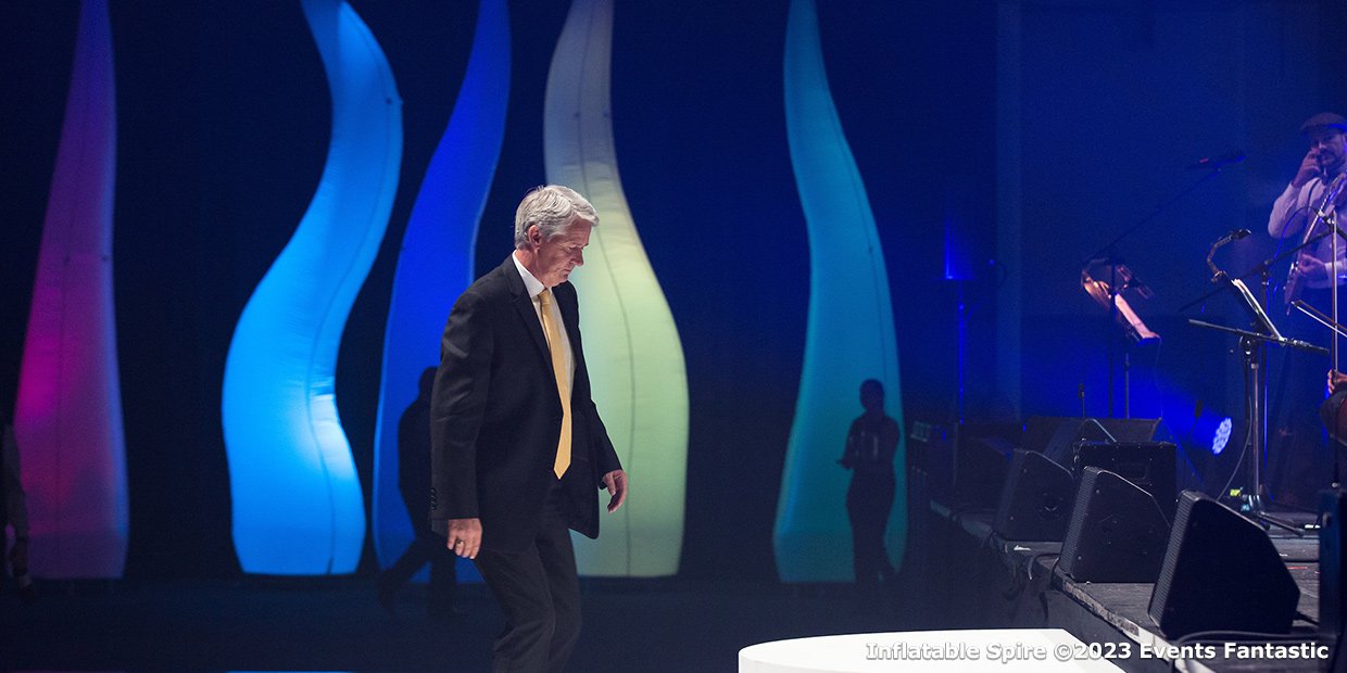 A man in a suit walks across a stage with colorful, vertically elongated lighting structures resembling spires in the background. Musicians are visible on the right side; the scene appears to be a live event.