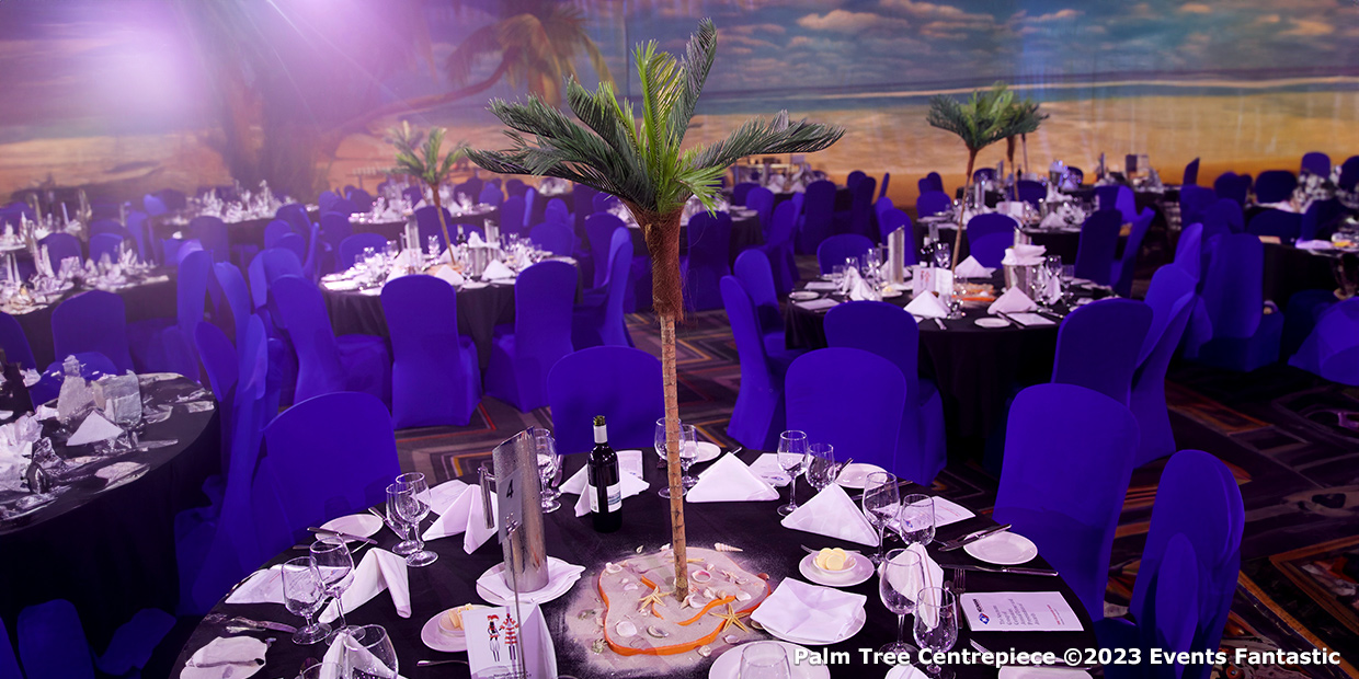 A banquet hall with multiple round tables covered in white linens and surrounded by purple chairs. Each table showcases a stunning palm tree centrepiece. The background beautifully resembles a beach scene, adding to the tropical ambiance.