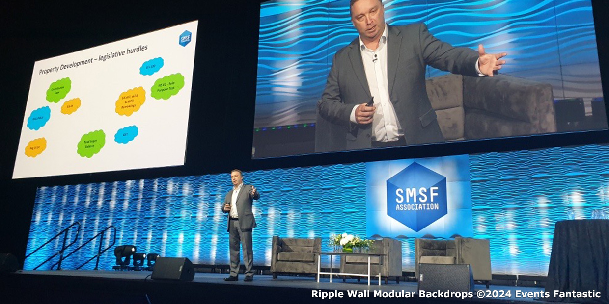 Presenter speaking on stage with a large screen displaying a slide on "Property Development - Legislative Hurdles." The background, featuring ripple wall modular panels, prominently highlights the large blue SMSF Association logo.