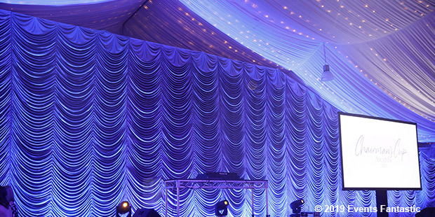 A stage setup with draped curtains and a large screen displaying text under a starry, chandelier-lit ceiling. © 2019 Events Fantastic is noted in the image corner.