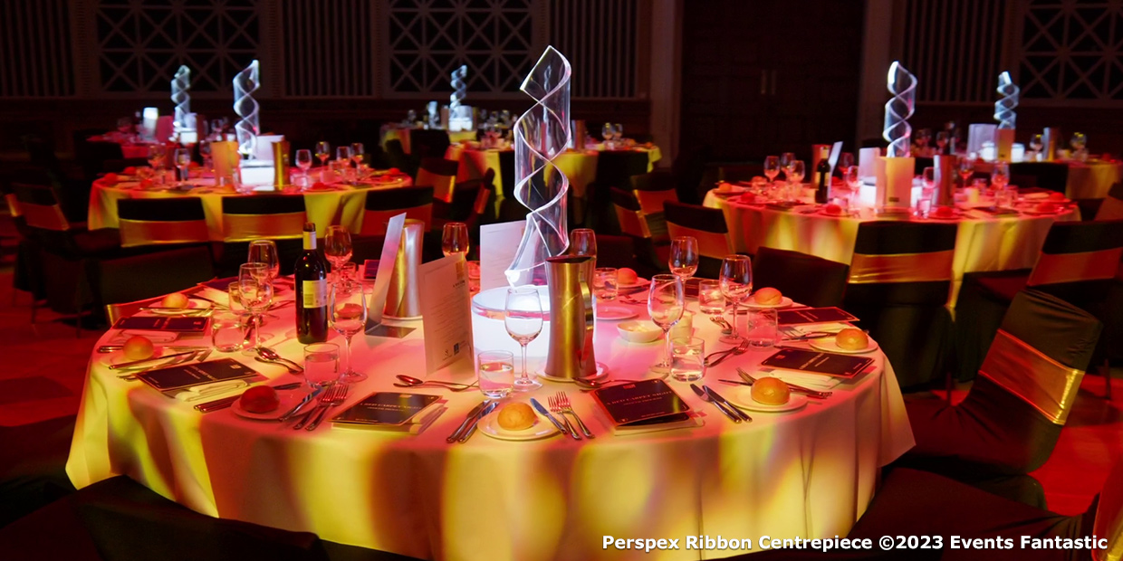 A formal event setting with round tables, illuminated Perspex Ribbon Centrepieces, neatly arranged utensils, plates, wine glasses, and folded napkins, all enveloped in a dim, warm lighting ambiance.