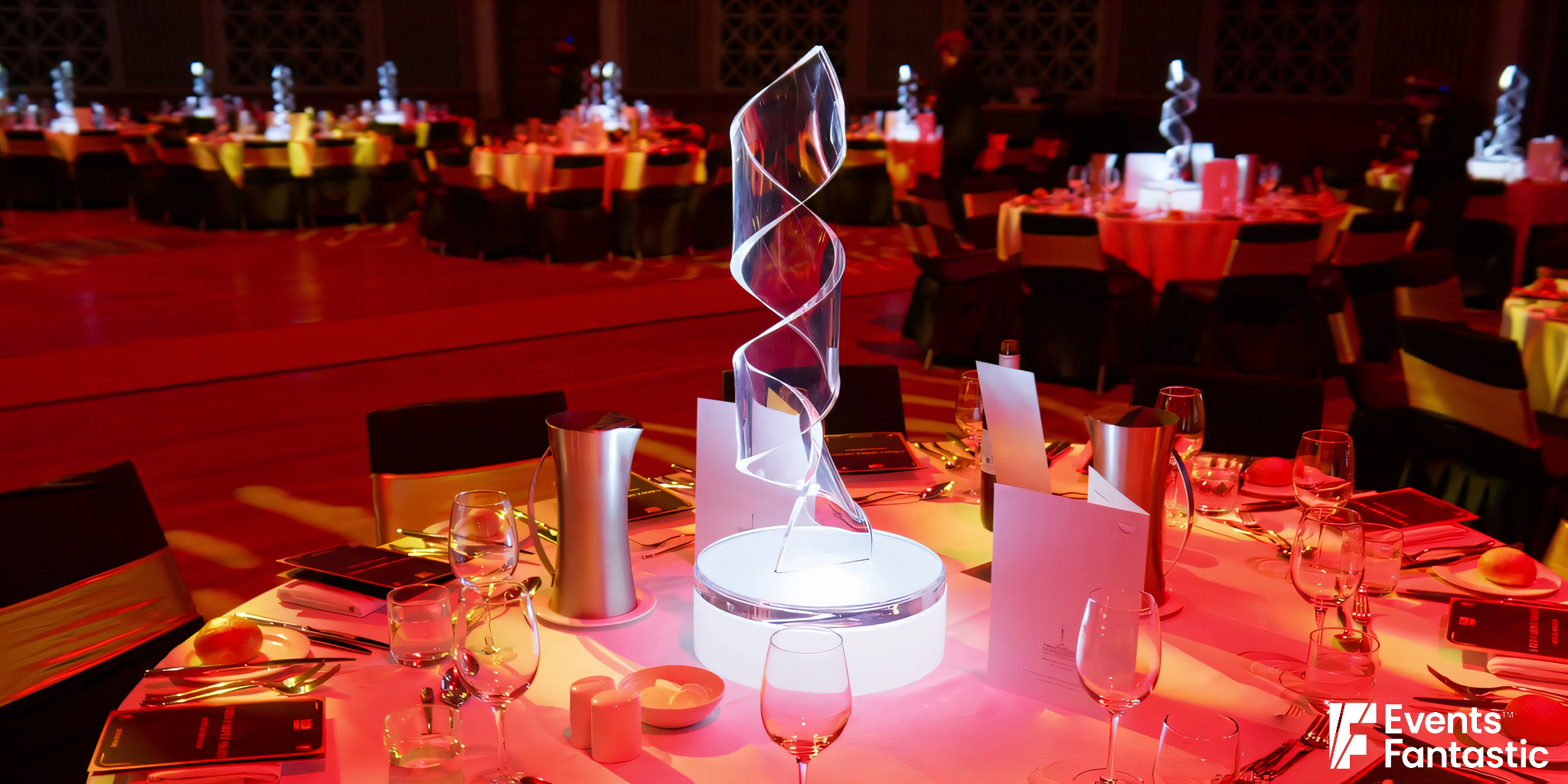 A decorated banquet hall with round tables set up with glassware, place settings, and tall, spiral Perspex ribbon centerpieces illuminated from below.