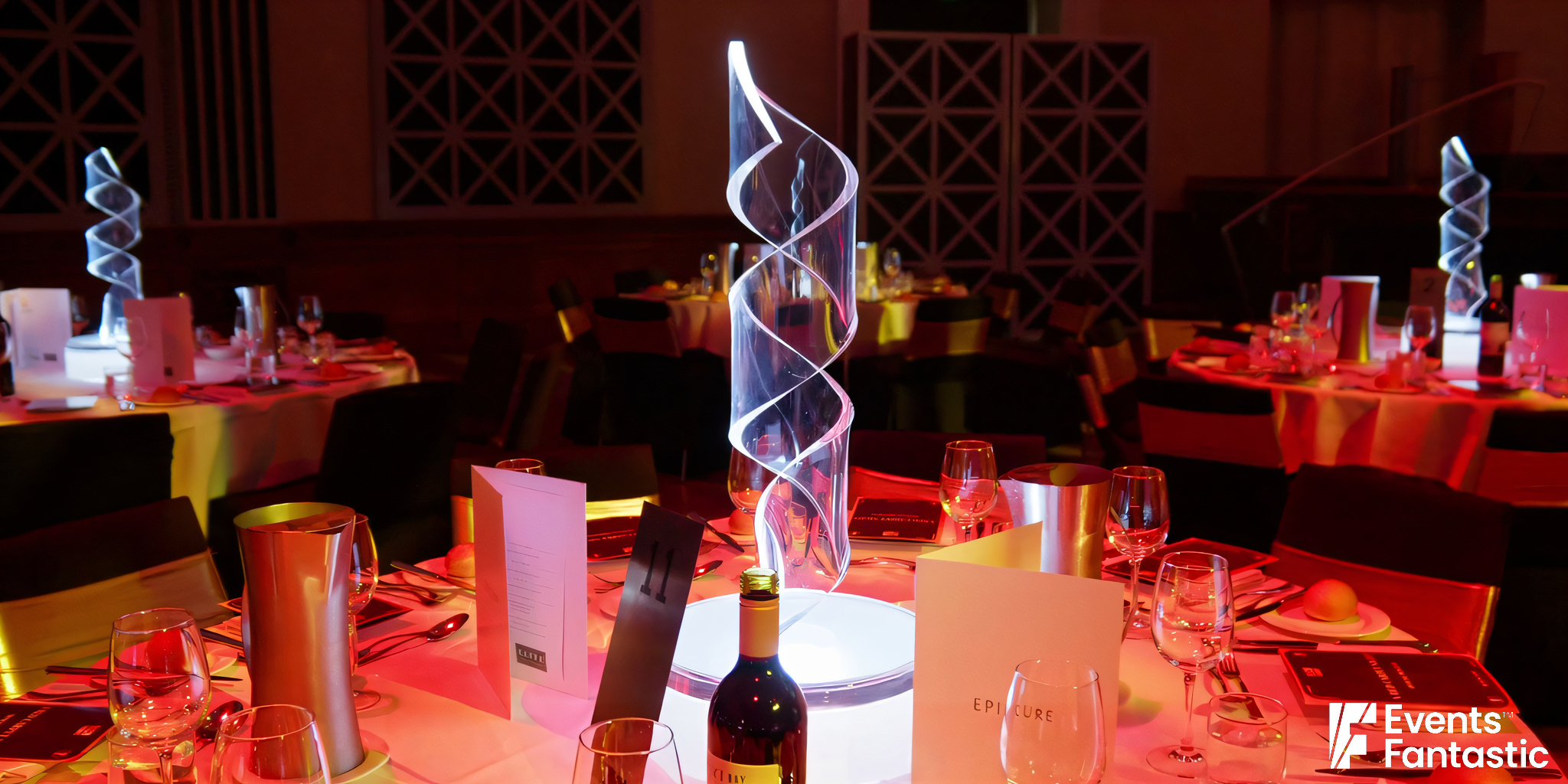 A formal dining setup featuring an illuminated Perspex Ribbon Centrepiece on round tables with menus, wine glasses, and a bottle of wine.