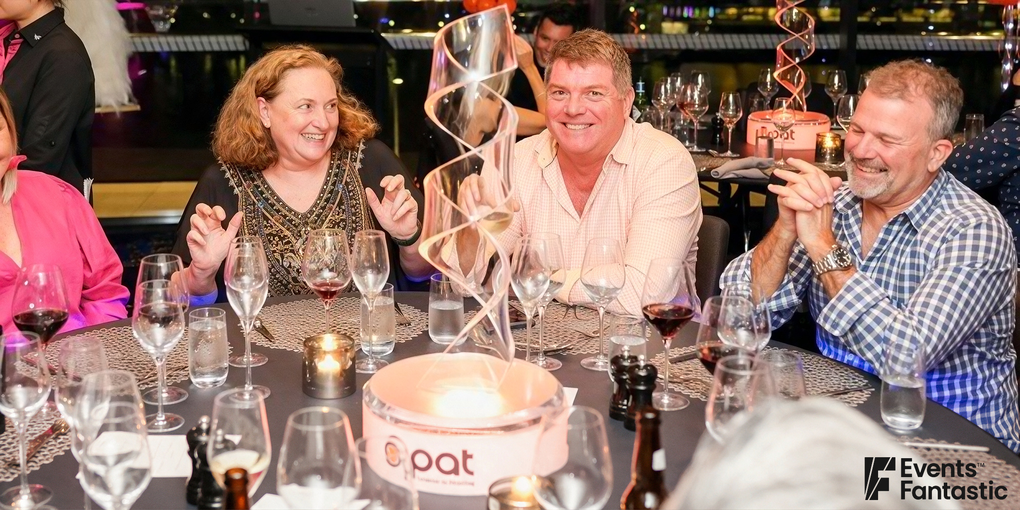 Four people sit at a round table covered with empty wine glasses, a Perspex ribbon centerpiece, and a lit candle in a dimly lit setting.