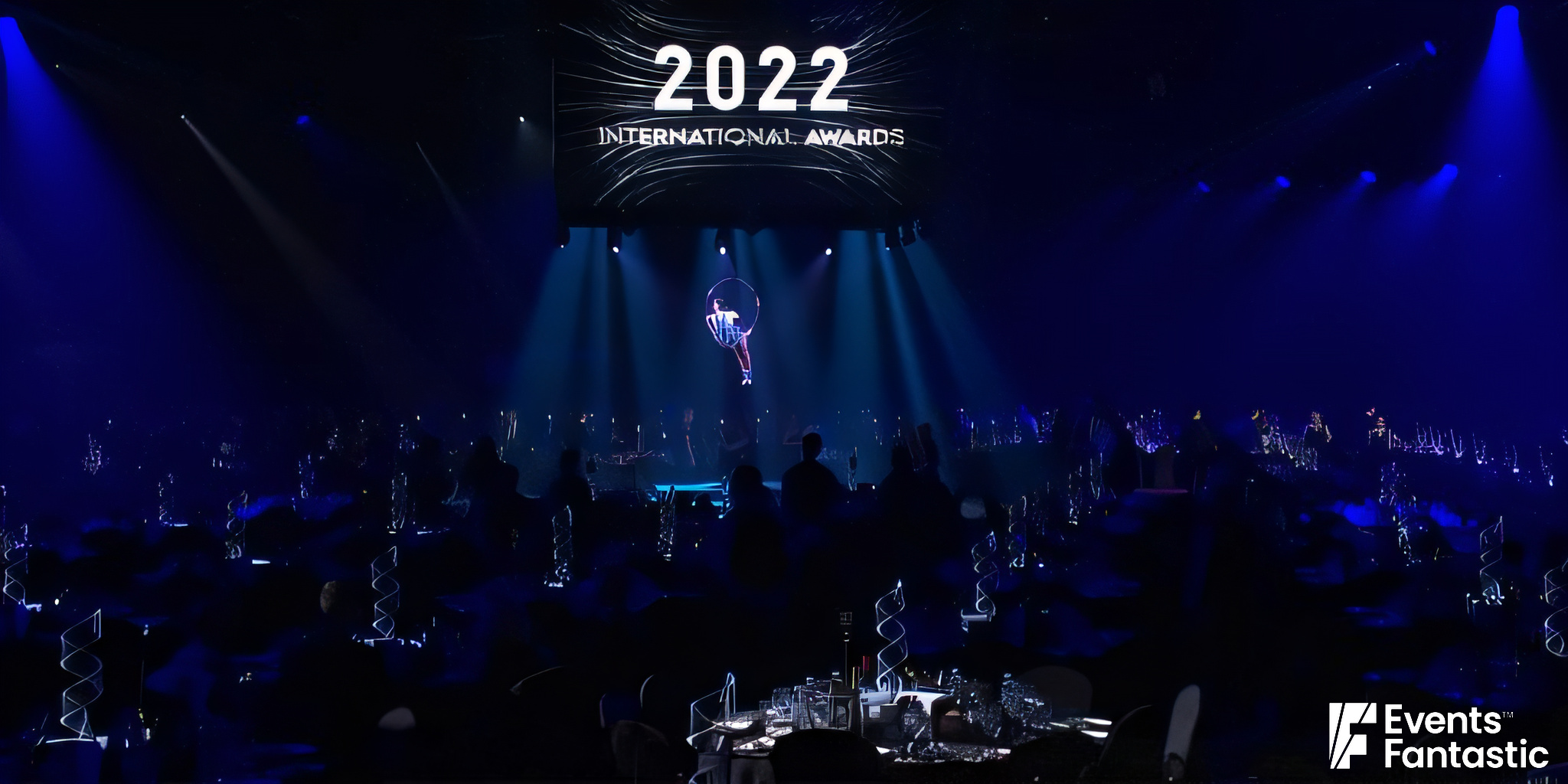 A stage with "2022 International Awards" displayed on a screen, featuring a performer suspended in a hoop, surrounded by a dimly lit audience. The scene is elevated by an elegant Perspex ribbon centrepiece adding a touch of modern sophistication.