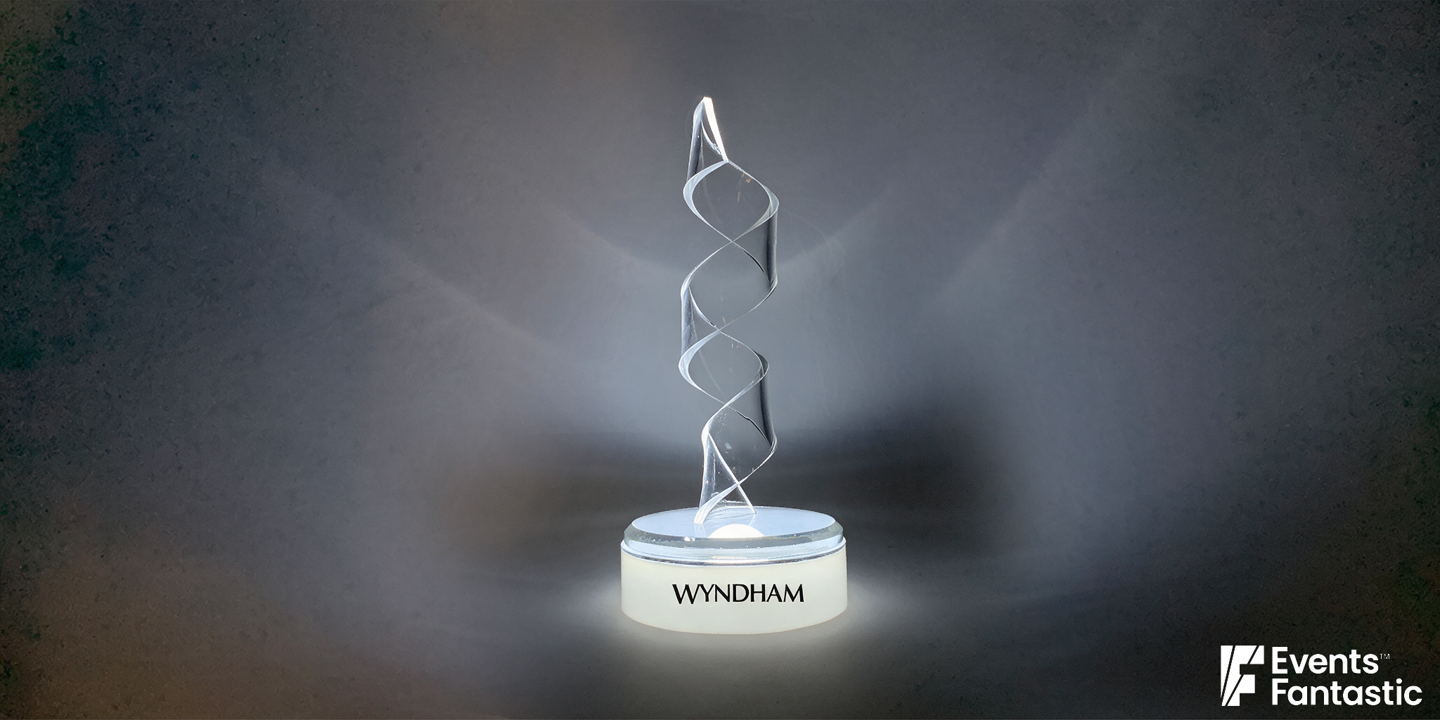 A clear, spiral-shaped award trophy on a circular base with the word "Wyndham" on it. The Perspex ribbon design adds a touch of elegance. The background is dark, accentuating the glowing edges of the trophy. © 2019 Events Fantastic.