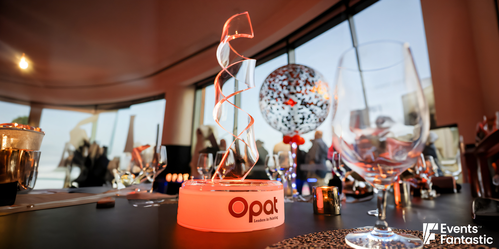 A ribbon-shaped Perspex centerpiece with the word "Opat" sits on a table surrounded by glassware in a brightly lit room.