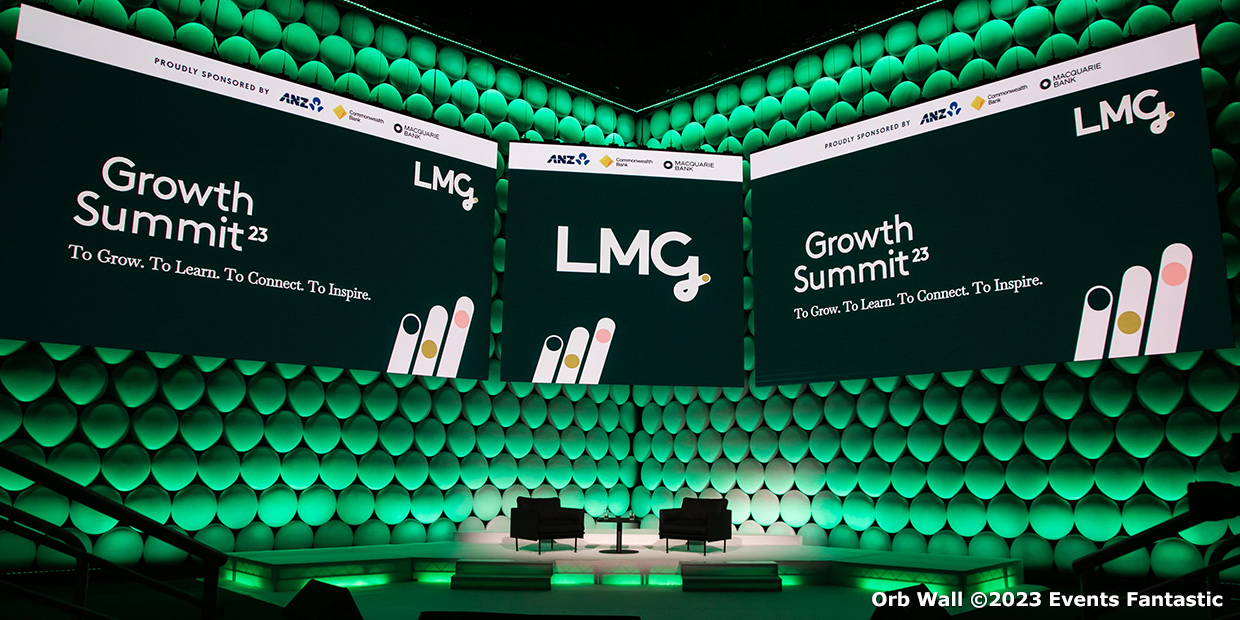 A stage setup for the Growth Summit '23 with large screens displaying event information and two chairs. The background features a textured green orb wall, adding a modern touch. Event sponsored by ANZ, Deloitte, and Optus.