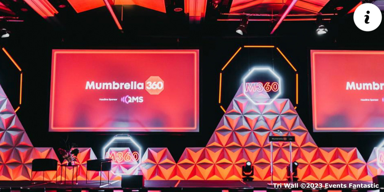 A stage setup for the Mumbrella 360 event featuring geometric backgrounds crafted from Tri Wall Modular Panels, vibrant red lighting, and large screens displaying the event name and sponsors. A podium and microphones are visible.