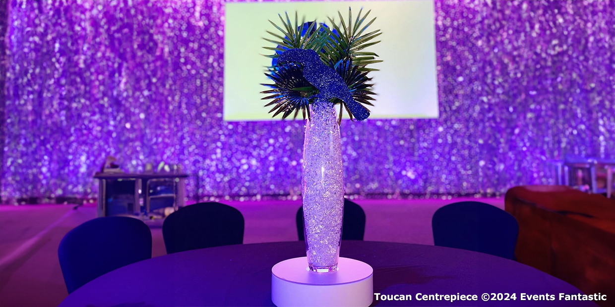 A tall, illuminated Havana Nights Toucan Centrepiece with sparkling decorations on a table, set against a purple-lit background.
