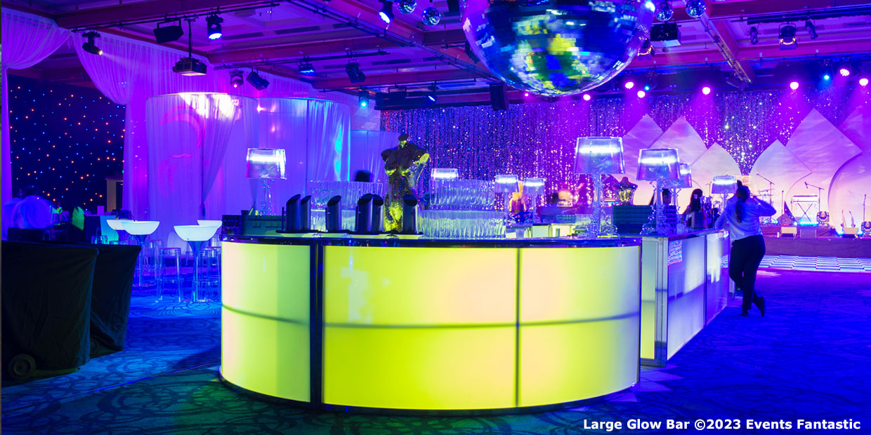 A large illuminated glow bar with glowing panels and a mirrored disco ball hanging above, set in a dimly lit room with decorative elements and several people.
