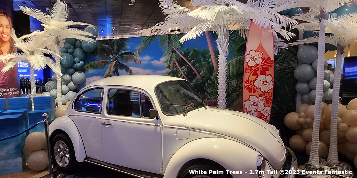 A white vintage car is displayed indoors, surrounded by Gold Palm Tree Props and balloons in various shades of blue, with a tropical backdrop featuring palm trees and a beach.