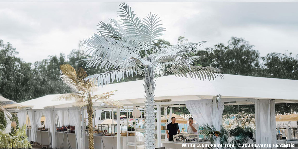 A large artificial white palm tree stands in front of a white event tent. Two people are visible under the tent, with trees in the background. Text at the bottom reads, "White 3.6m Palm Tree ©2024 Events Fantastic." For added elegance, consider our Gold Palm Tree Prop.