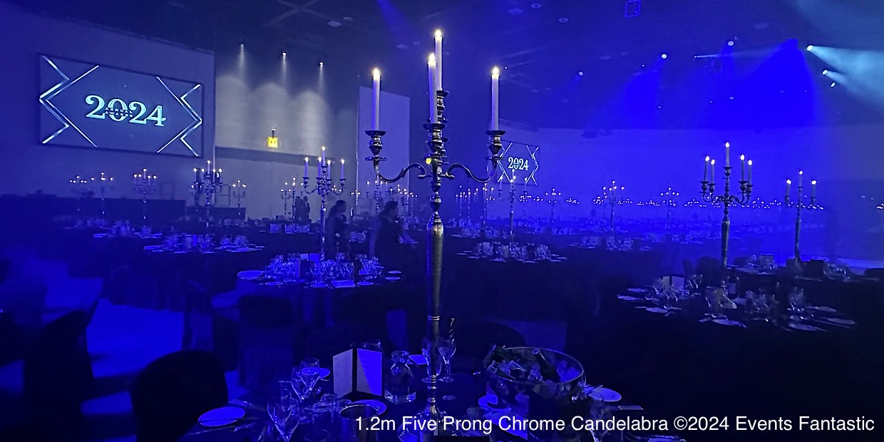 Dimly lit banquet hall set for an event with tall, chrome 5 prong candelabras and round tables, with "2024" displayed on a screen in the background.