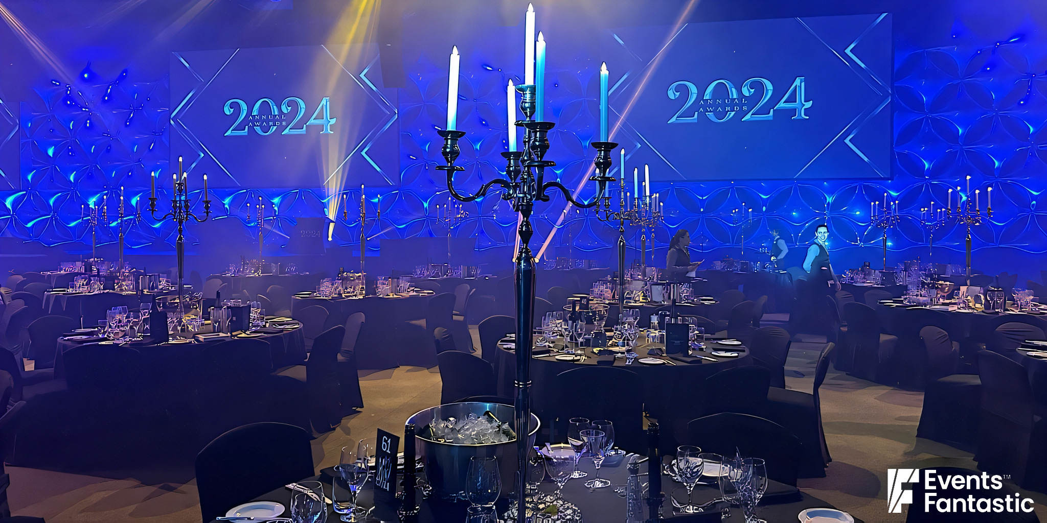 A large banquet hall is set for an event with round tables, elegant place settings, and chrome 5-prong candelabras. The large screens display "2024.