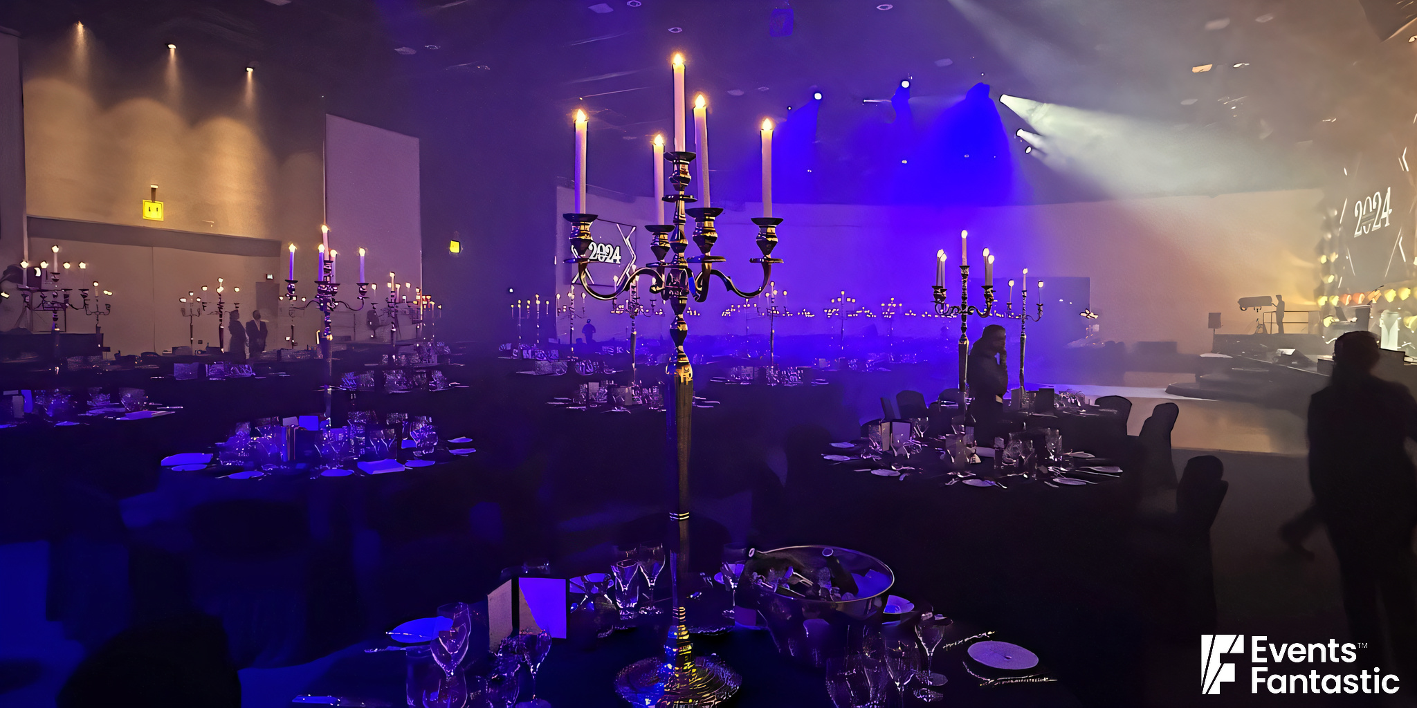 A dimly lit banquet hall with round tables set for an event. Tall chrome 5 prong candelabras with lit candles adorn each table. Blue lighting creates a moody atmosphere.