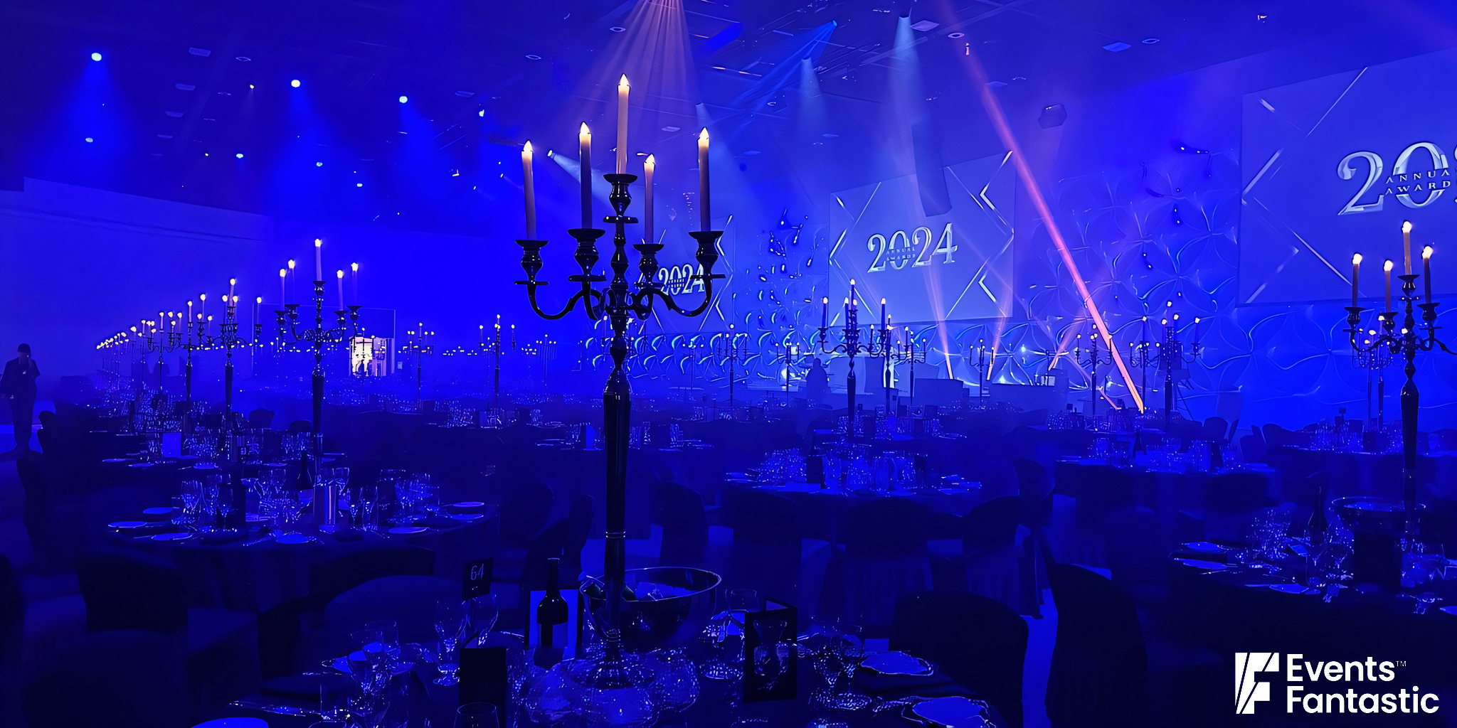 A dimly lit event hall with multiple tables set for a formal gathering. Tall chrome 5 prong candelabras adorn each table. A large screen displays “2024.” Blue lighting enhances the ambiance.