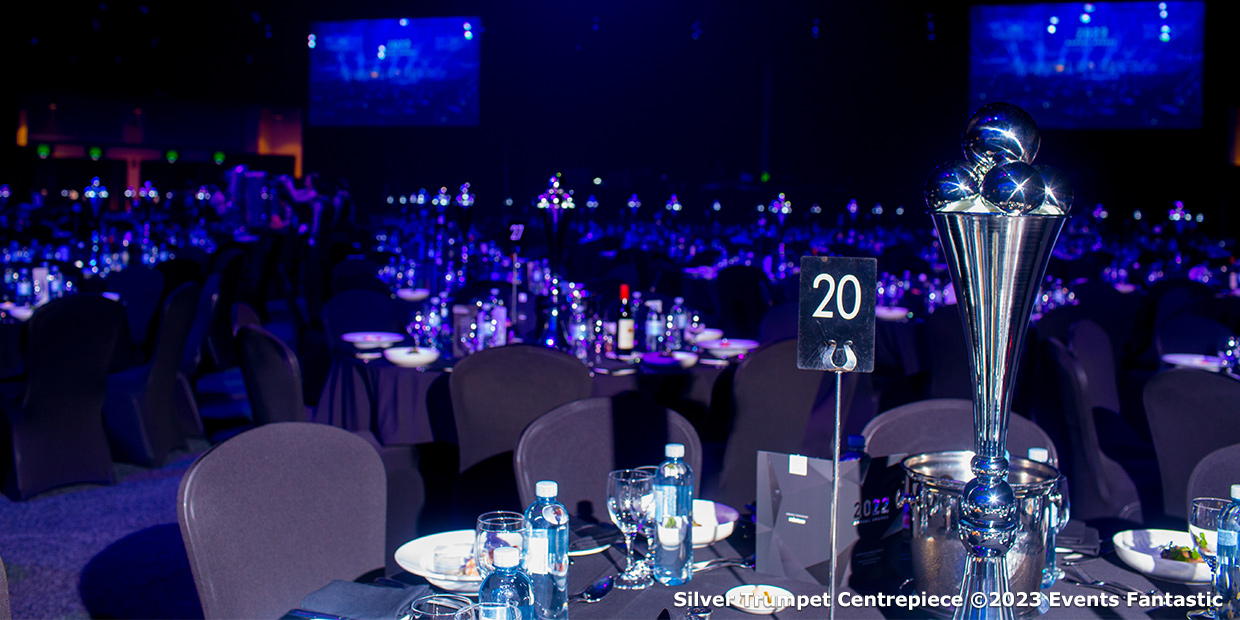 A dimly lit banquet hall features numerous round tables with black tablecloths, set with tableware. Each table has a centerpiece, including a Chrome 5 Prong Candelabra, and a number sign. A stage with blue lighting is visible in the background.