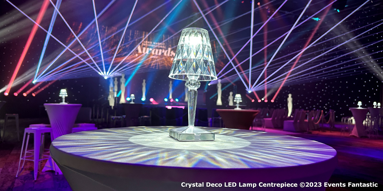 A decorated event venue with bright lights and tables featuring Crystal Deco LED Centrepieces. The backdrop displays "Awards" text. Caption reads: "Crystal Deco LED Lamp Centrepiece ©2023 Events Fantastic.