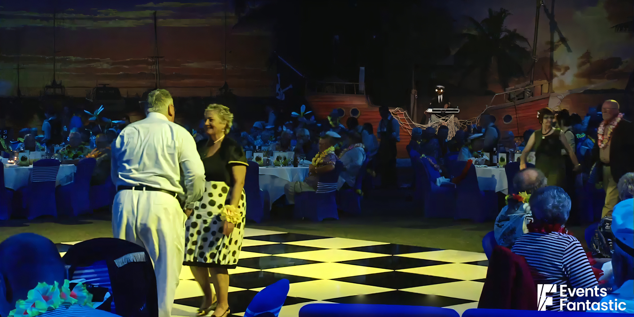 An older couple is dancing on a shiny black-and-white checkered dance floor at an event. Guests are seated at decorated tables in the background against a tropical-themed backdrop, showcasing the elegance of Black and White Shiny Dance Floor Hire.