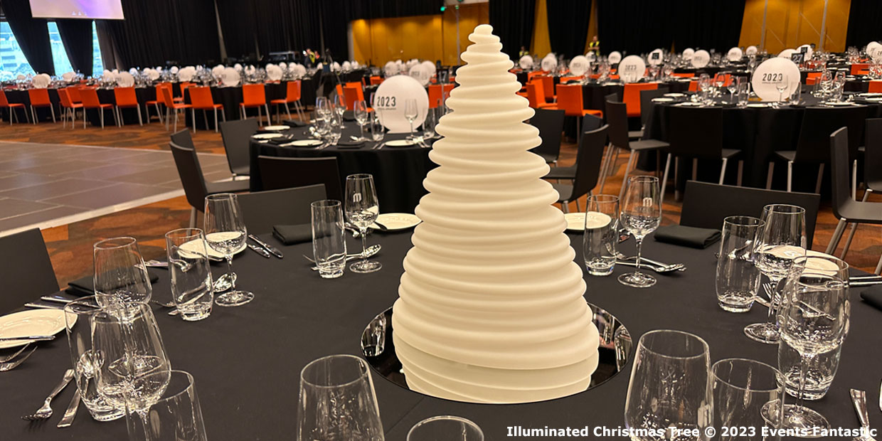 A white, conical sculpture resembling an abstract Christmas tree, with an illuminated glow, is the centerpiece on a dining table set with glassware and cutlery. The background includes numerous similarly set tables and orange chairs.