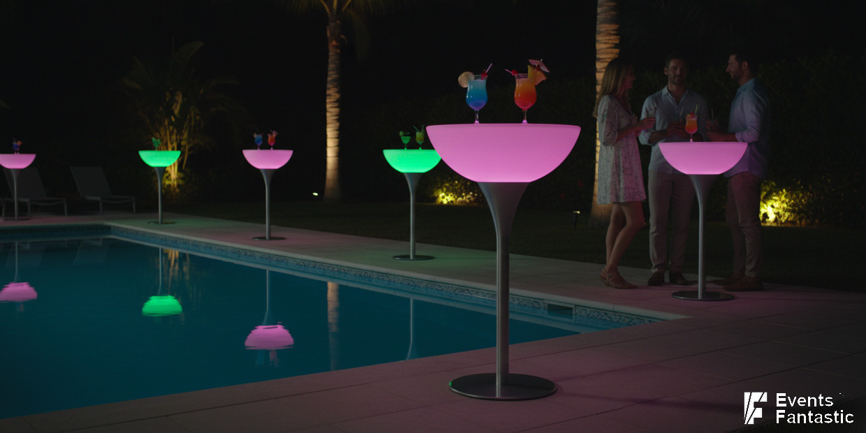 People stand by a pool at night, gathered around glow cocktail bar tables with a metal base, each holding vibrant drinks. Palm trees and soft outdoor lighting create an inviting atmosphere in the background.