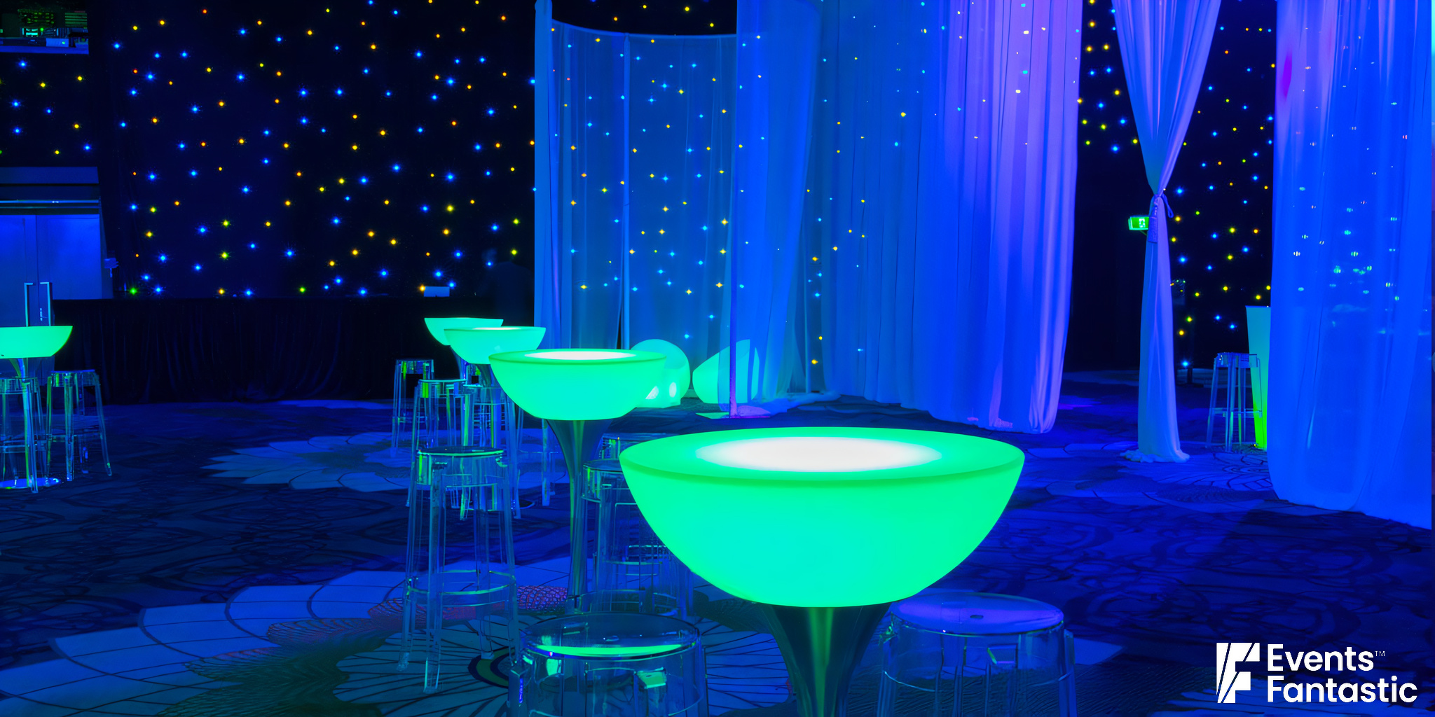 A room with illuminated glow cocktail tables featuring metal bases and transparent stools is set against a dark starry backdrop with curtain partitions.