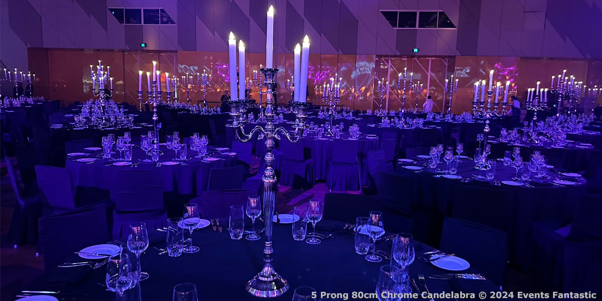 Dimly lit event hall with round tables set for a formal dinner, featuring chrome 5-prong candelabras with lit candles as elegant centerpieces.
