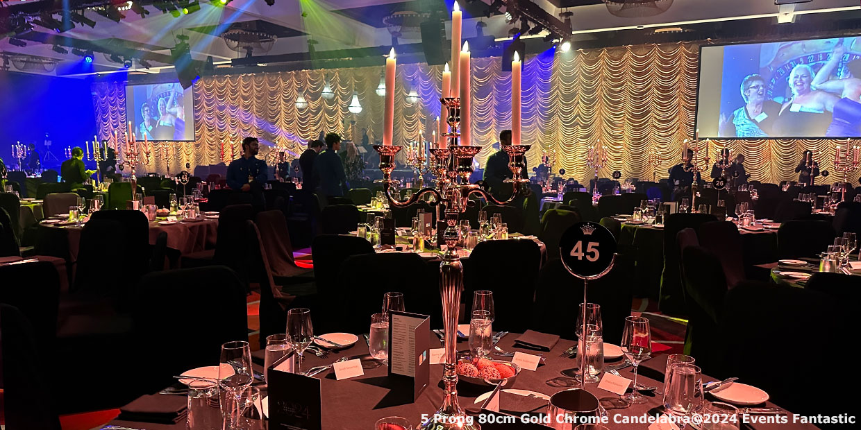 A banquet hall with elegantly set tables, chrome 5 prong candelabras, and a large stage with lighting effects. Two screens display images on either side. The table in the foreground is numbered 45.