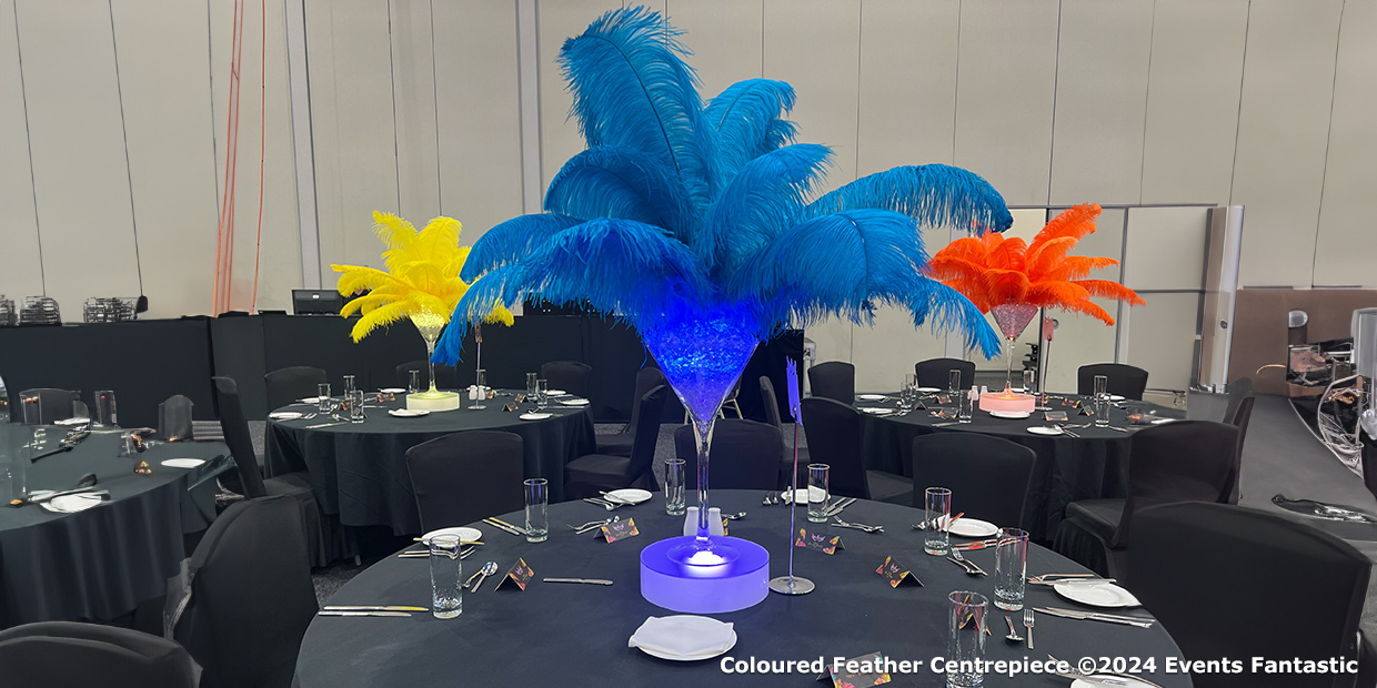 A banquet room with round tables, each adorned with Gatsby-inspired centerpieces made of bright-colored feathers in shades of blue, yellow, and orange. The tables are set with glassware and black cloths for a sophisticated touch.