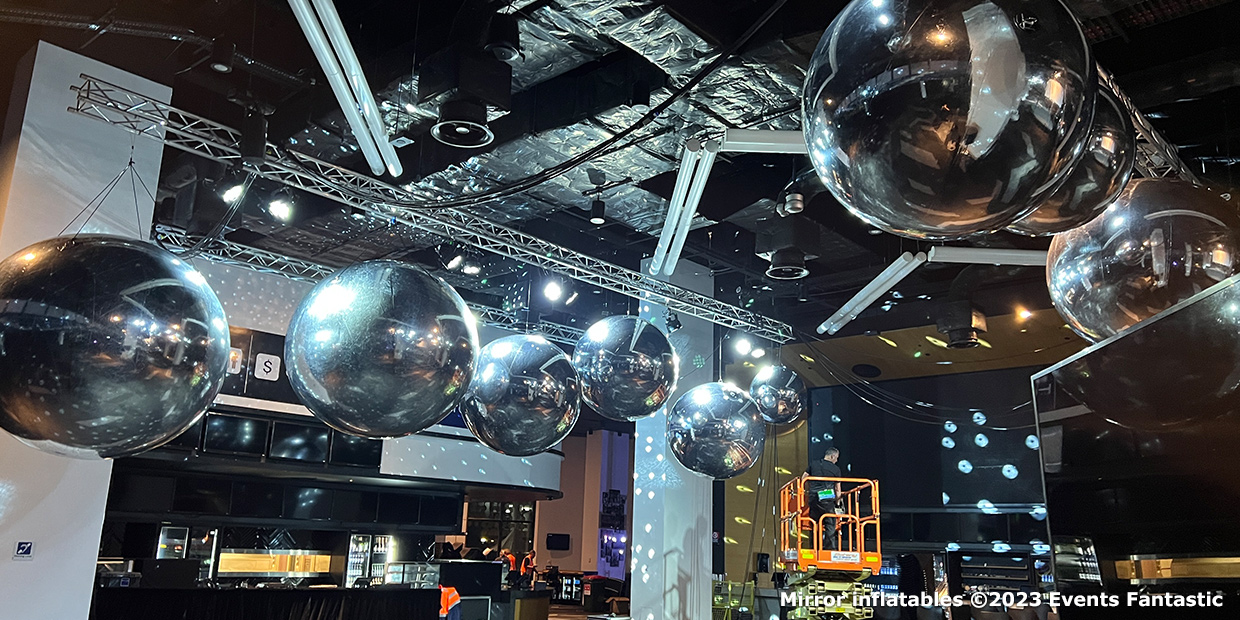 Large silver shiny mirror balls hanging from the ceiling in a modern event space. Stage lighting and equipment are visible in the background.
