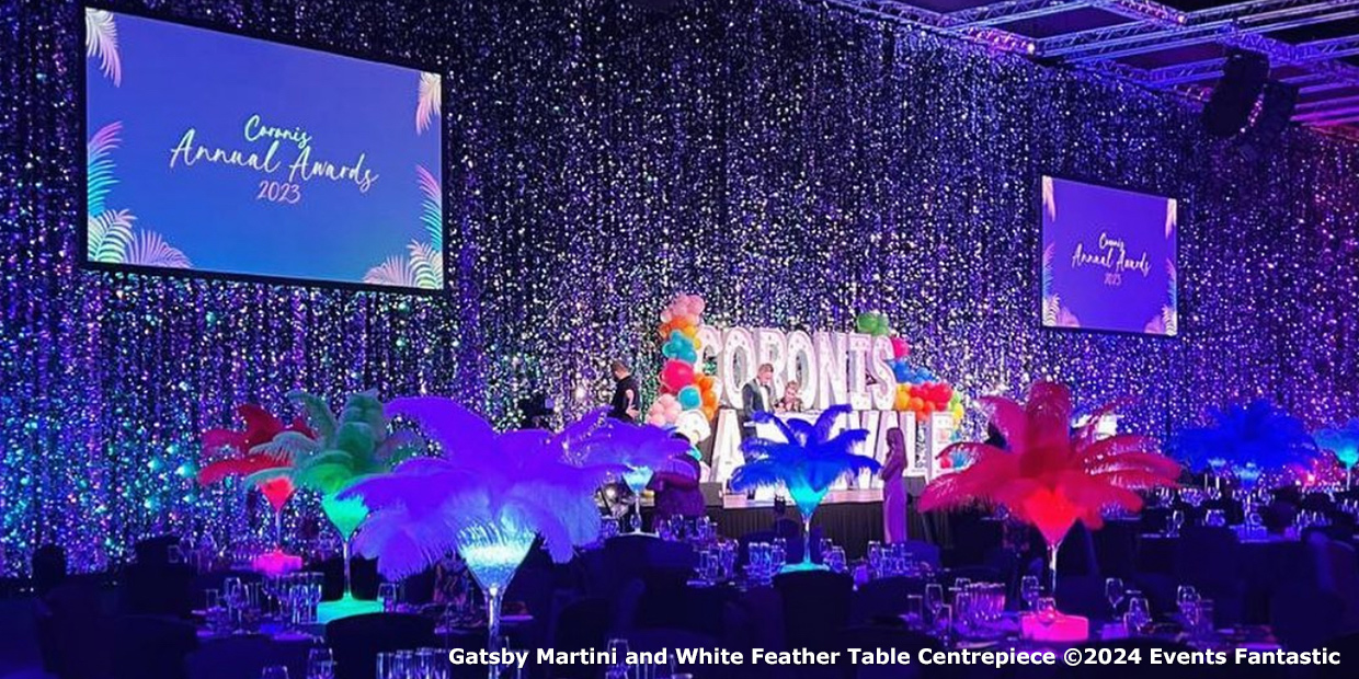 A stage with colorful decorations at an awards event. Two screens display "Coming Annual Awards 2023." Coloured feather centerpieces illuminate round dining tables in the foreground.