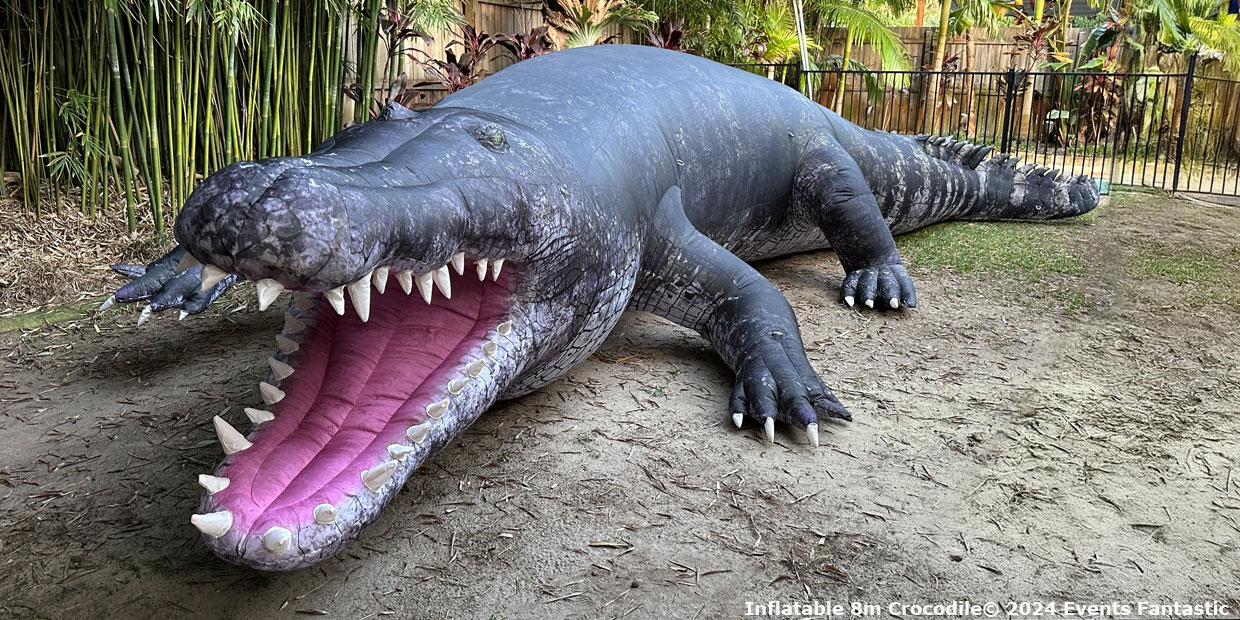 A large 8m inflatable crocodile rests on a dirt ground next to a fence. It has an open mouth with visible white teeth. Text at the bottom reads, "Inflatable 8m Crocodile © 2024 Events Fantastic.
