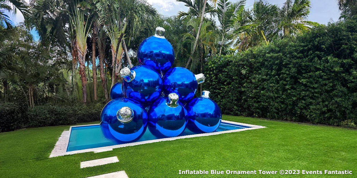 A large outdoor sculpture consisting of stacked blue inflatable ornaments forms a striking tower, surrounded by palm trees and greenery. Text in the bottom right corner reads "Inflatable Blue Ornament Tower ©2023 Events Fantastic.