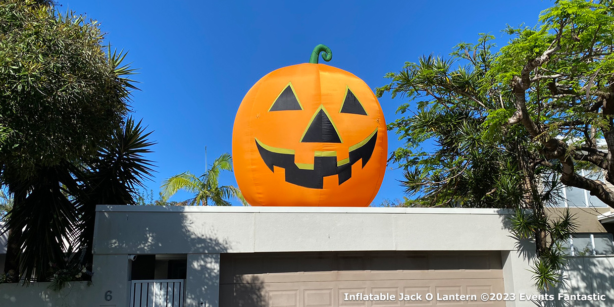 A large inflatable Jack O' Lantern with a smiling face sits on top of a garage, surrounded by trees and clear blue sky, creating a festive atmosphere that could rival any Inflatable Santa.