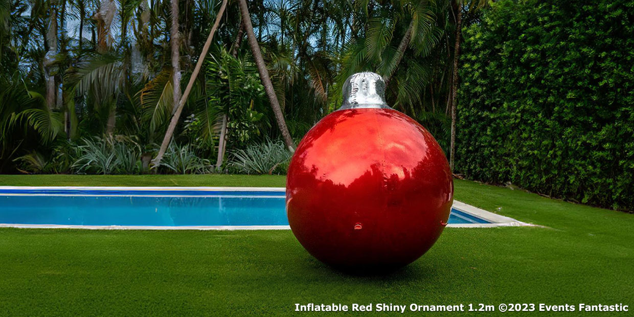 A large, red, shiny inflatable ornament stands proudly on a green lawn next to a swimming pool surrounded by trees. Text in the corner reads "Inflatable Red Shiny Ornament 1.2m ©2023 Events Fantastic.