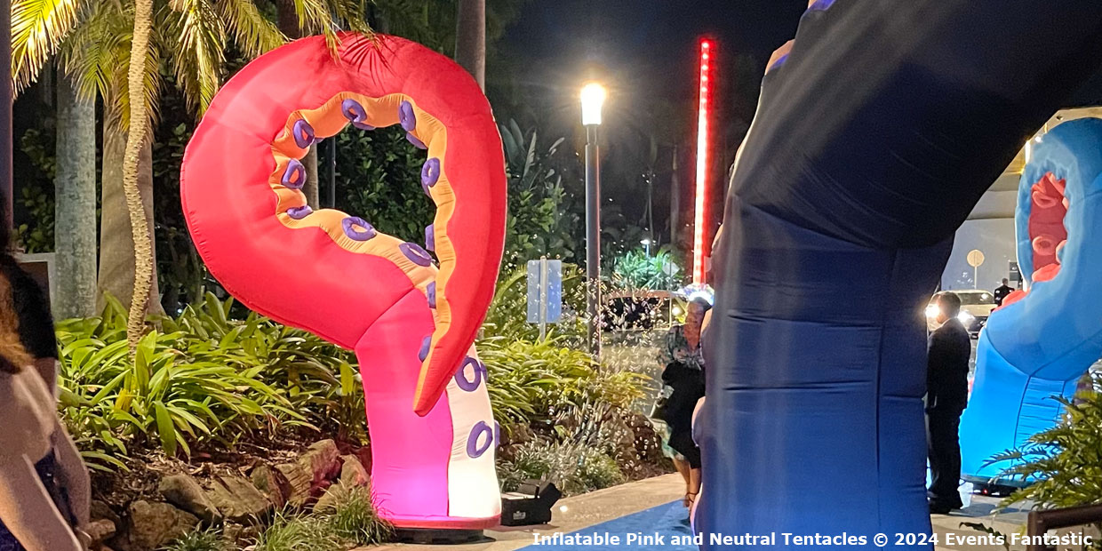 Inflatable pink and neutral tentacles are displayed outdoors at night among plants and people, creating an enchanting atmosphere. A narrow pathway is visible leading through the setup.