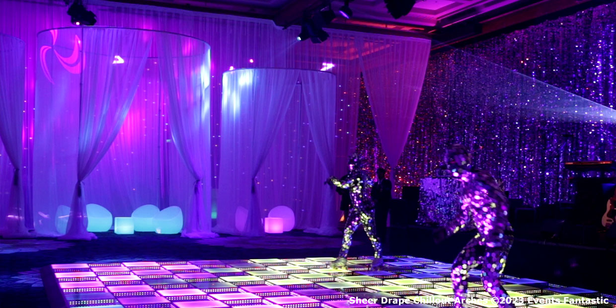 Two performers in reflective suits dance on a lit floor in a room with sheer drapes and vibrant purple lighting. The drapes, hanging 3m high, create an ethereal atmosphere as the dancers move gracefully in circular patterns.