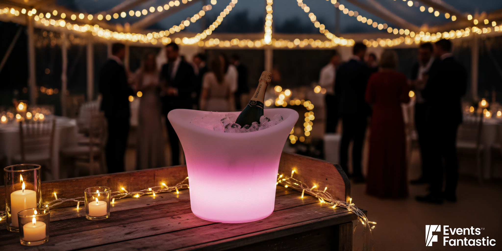 An illuminated ice bucket with a bottle of champagne sits on a decorated table at an evening event, as guests mingle under string lights in the background.