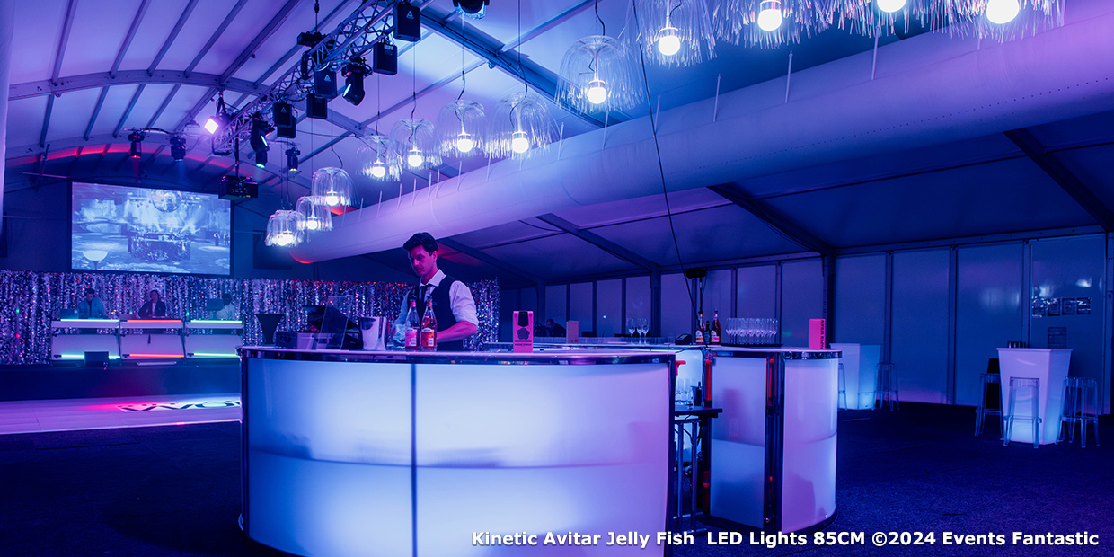 A bartender stands behind a glowing circular bar in a large, modern event space, illuminated by blue and purple lighting and hanging 85cm Jelly Fish LED Lights that create a dazzling underwater ambiance.