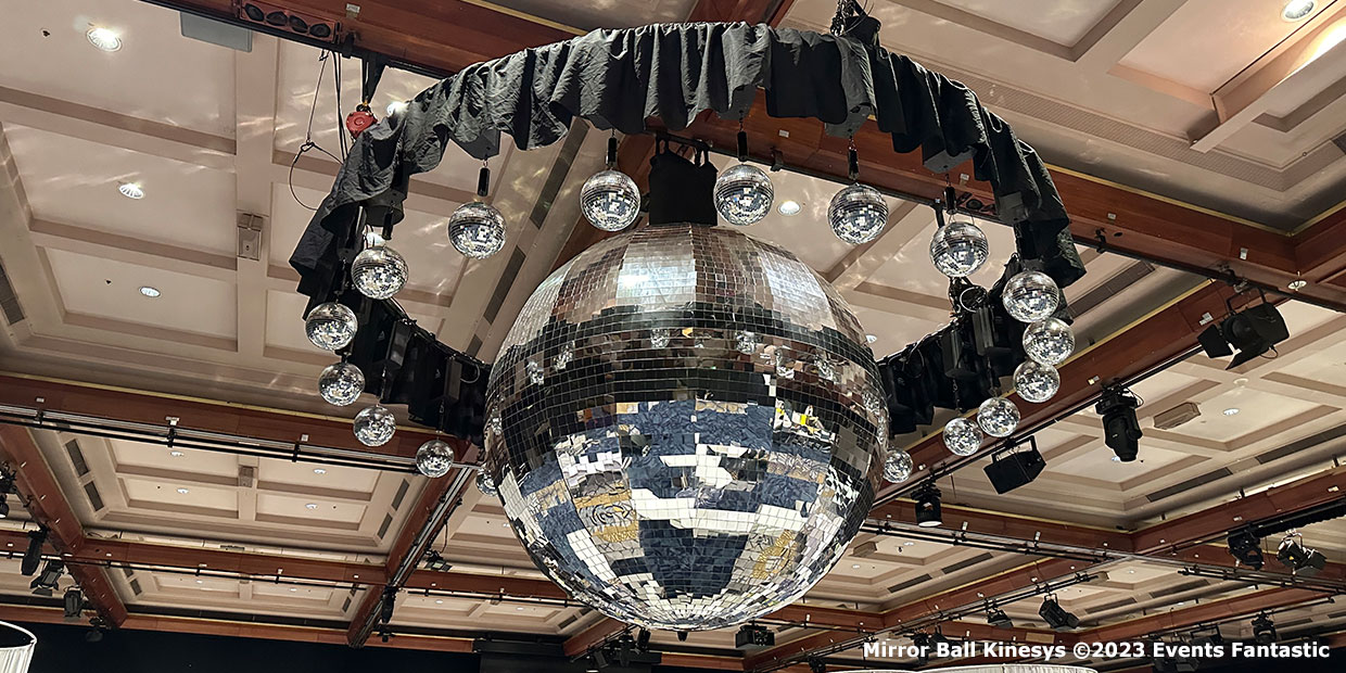 A large mirrored disco ball, accented by 25cm Kinetic Mirror Balls and smaller globe lights, hangs from a ceiling with exposed beams in an event venue.