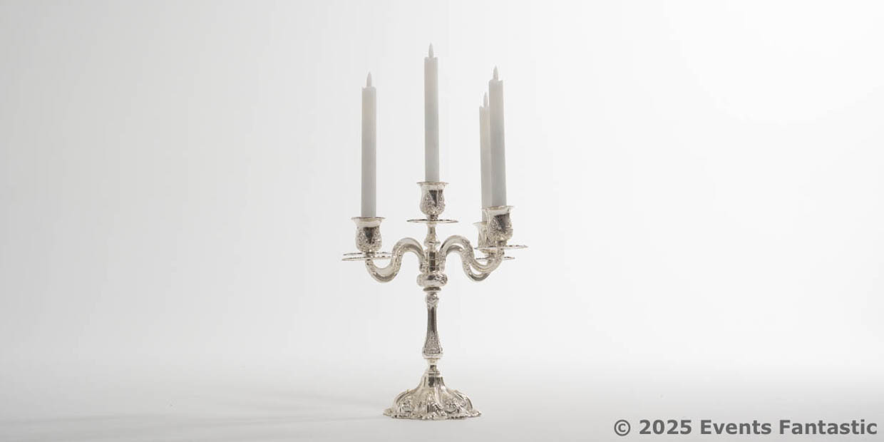 A Chrome 5 Prong Candelabra holds five unlit white taper candles, elegantly displayed against a plain white background.